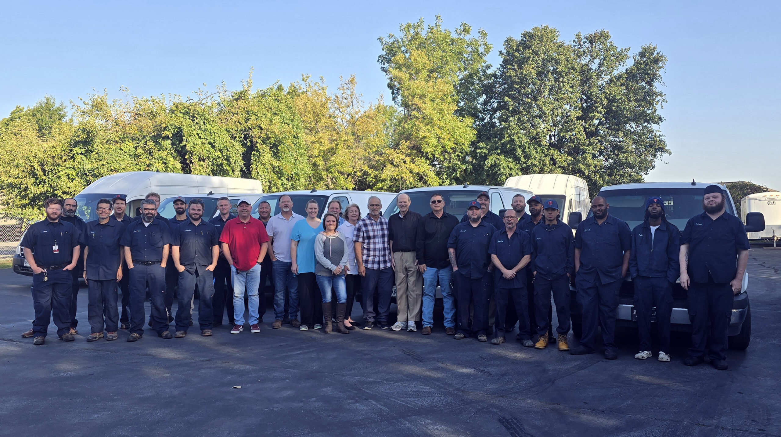 Photo of over 30 employees standing outside in a black top parking light in front of their white fleet of vans and trucks.