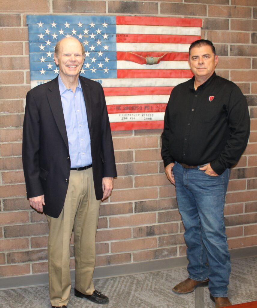 Left to right: Owners Mark Simmons and Todd Thompson. Mark is wearing a nice black jacket with light blue collared shirt with tan pants and black belt with shiny loafer style shoes with tassels on the front of the shoes. Todd is wearing a long sleeved collared black ironed shirt with the red Allstate Fire Midwest shield logo. The american flag the KCMO Fire Department gave them is on the wall in the background hung on a multi-red brick wall in their conference room.